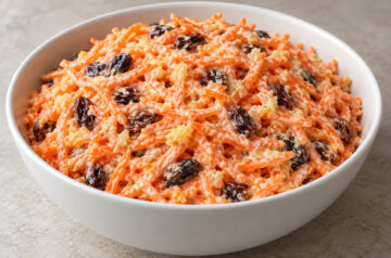 AI illustration of shredded carrot, raisin, and pineapple salad with creamy dressing in a white bowl.