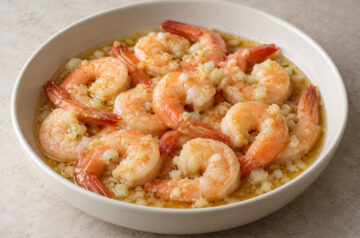 AI illustration of tail-on prawns in golden oil with chopped garlic and pale onion pieces in a white bowl.
